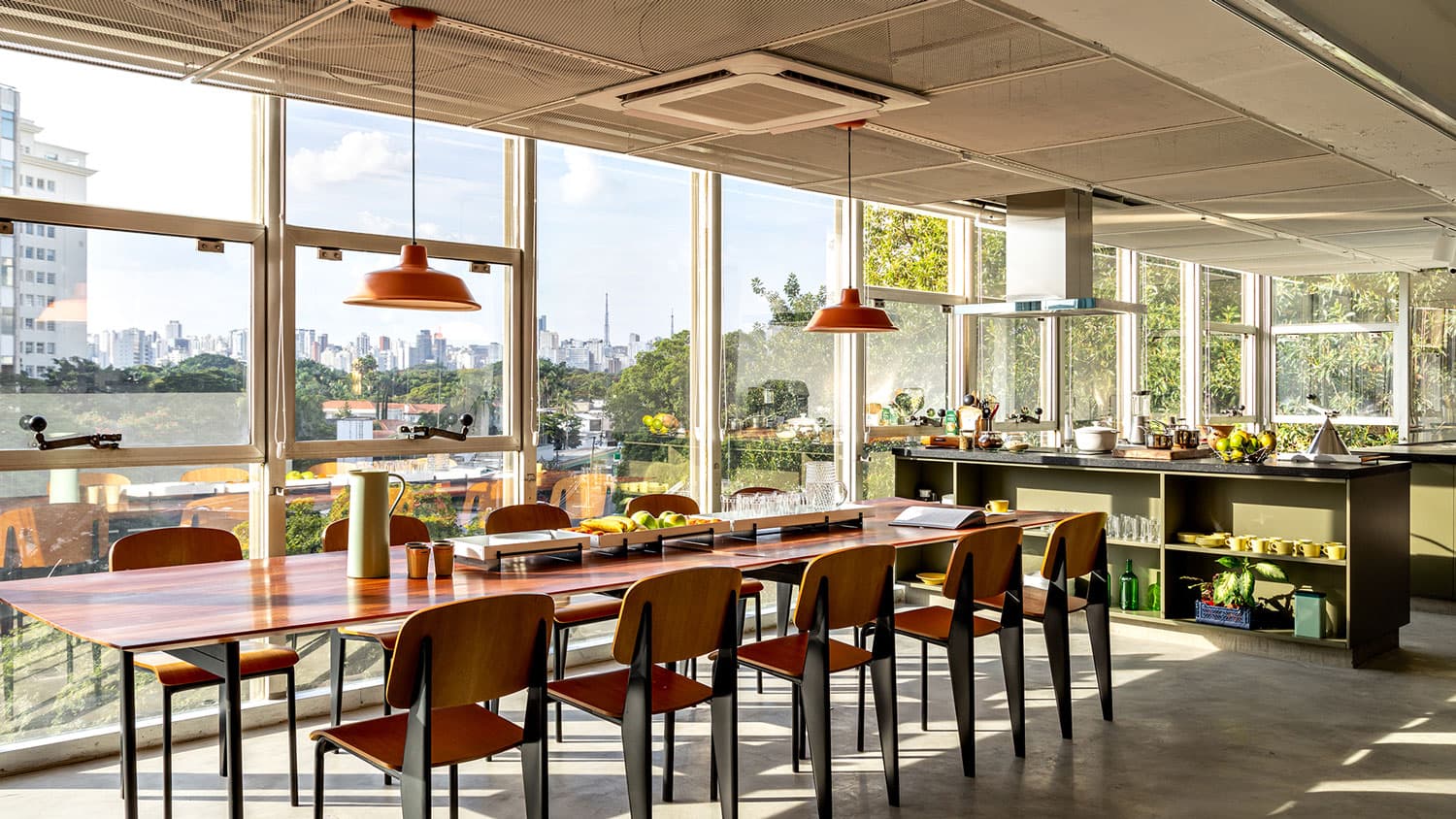 Refeitório do Bioma com mesa comunitária de madeira, luminárias laranja e vista de São Paulo